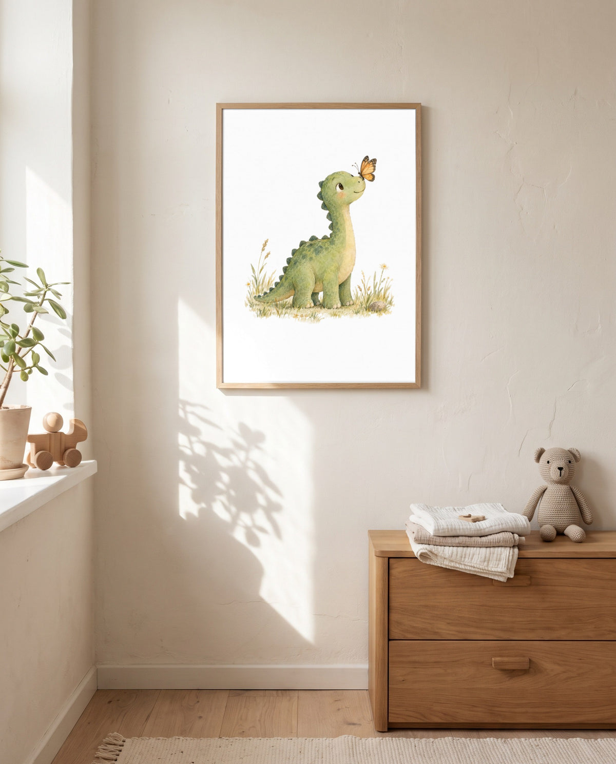 Gerahmtes Poster mit grünem Dinosaurier und Schmetterling an einer hellen Wand neben einem Fenster; darunter eine Holzkommode mit gefalteten Textilien und einem Stoffbären.
