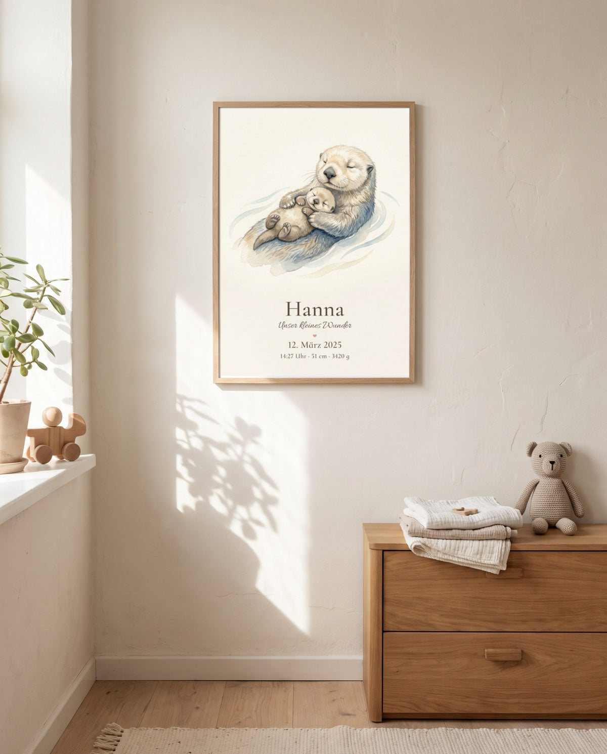 Otter mit schlafendem Jungtier als Poster im Holzrahmen über einer Holzkommode mit gefalteten Textilien und Stoffbär; links ein Fenster mit Pflanze und Holzspielzeug.
