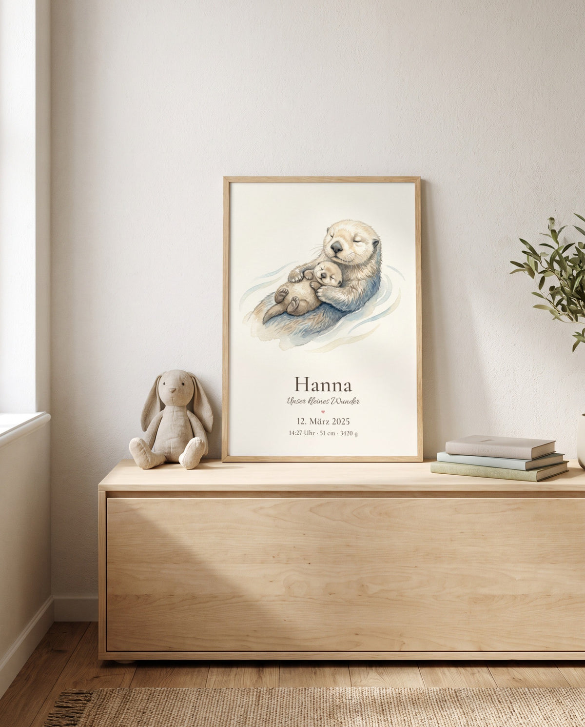 Gerahmtes Poster mit großem Otter und Jungtier auf einer niedrigen Holzkommode, daneben ein Stoffhase und ein Bücherstapel; seitlich sind Fenster und Pflanzenzweige sichtbar.
