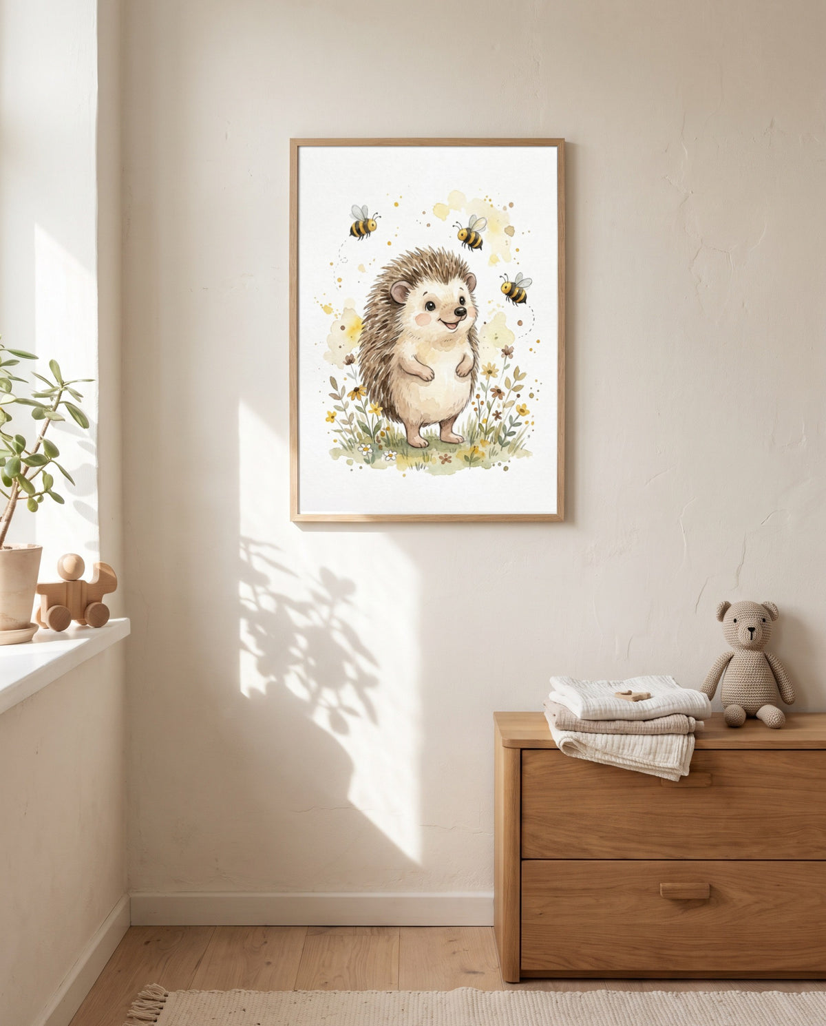 Igel-und-Bienen-Poster im Holzrahmen an einer sonnigen Wand über einer Holzkommode mit gefalteten Textilien und Stoffbär; links ein Fenster mit Pflanze und Holzspielzeug.