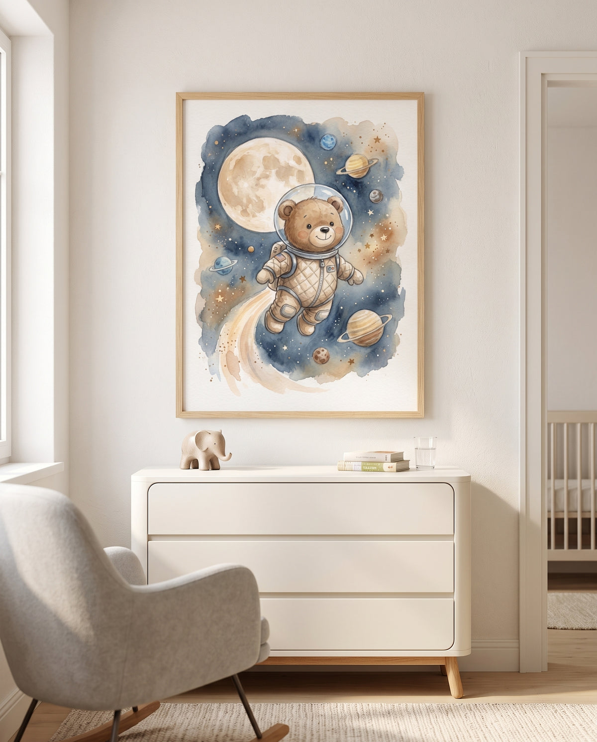 Poster mit einem Bären als Astronaut zwischen Mond, Sternen und Planeten im Holzrahmen über einer cremefarbenen Kommode mit Elefantenfigur und Büchern. Links steht ein grauer Polstersessel am Fenster, rechts ist durch eine offene Tür ein Gitterbett​