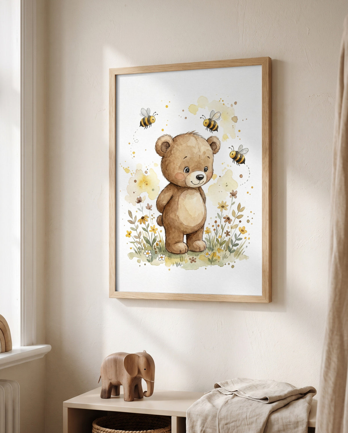 Poster mit Bär, drei Bienen und Blumen im hellen Holzrahmen neben einem Fenster, darunter ein niedriges Regal mit Holzelefant, Korb und hellem Stoff.
