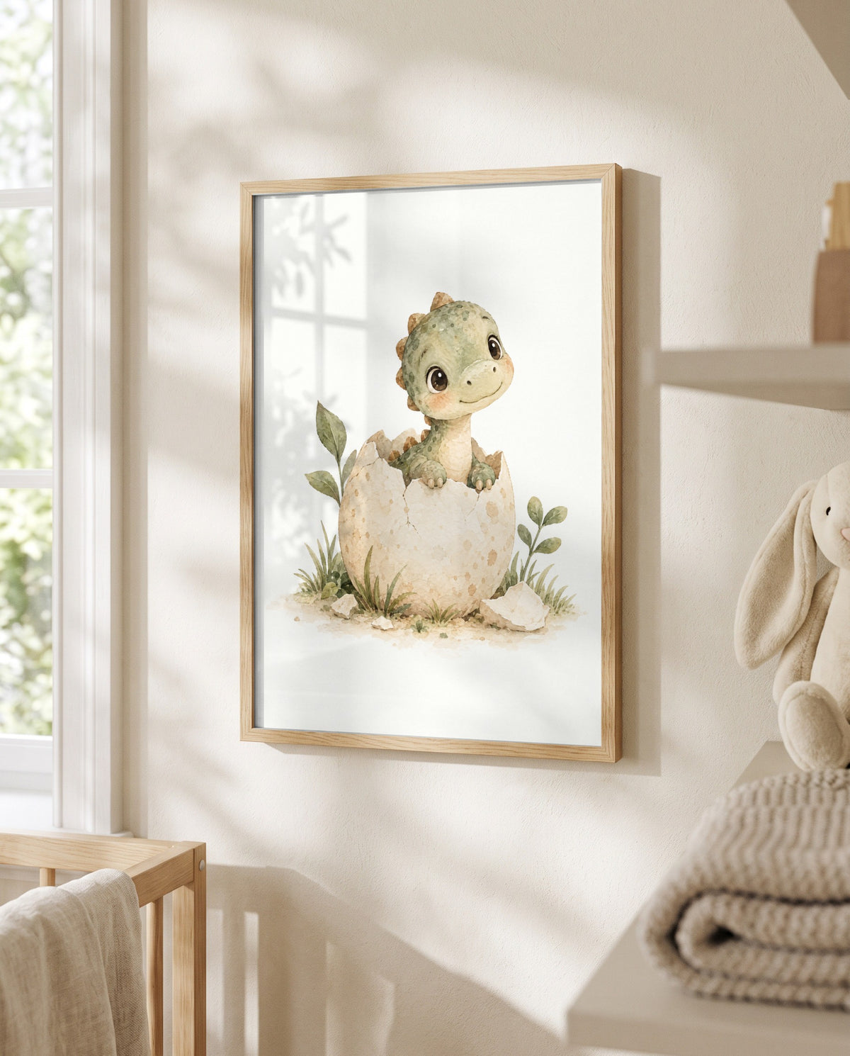 Dino-im-Ei-Poster im hellen Holzrahmen neben einem Fenster mit Licht- und Schattenmustern. Unten links ist ein Holzgitter mit übergelegtem Tuch sichtbar, rechts liegen gefaltete Textilien unter einem teilweise sichtbaren Stoffhasen.