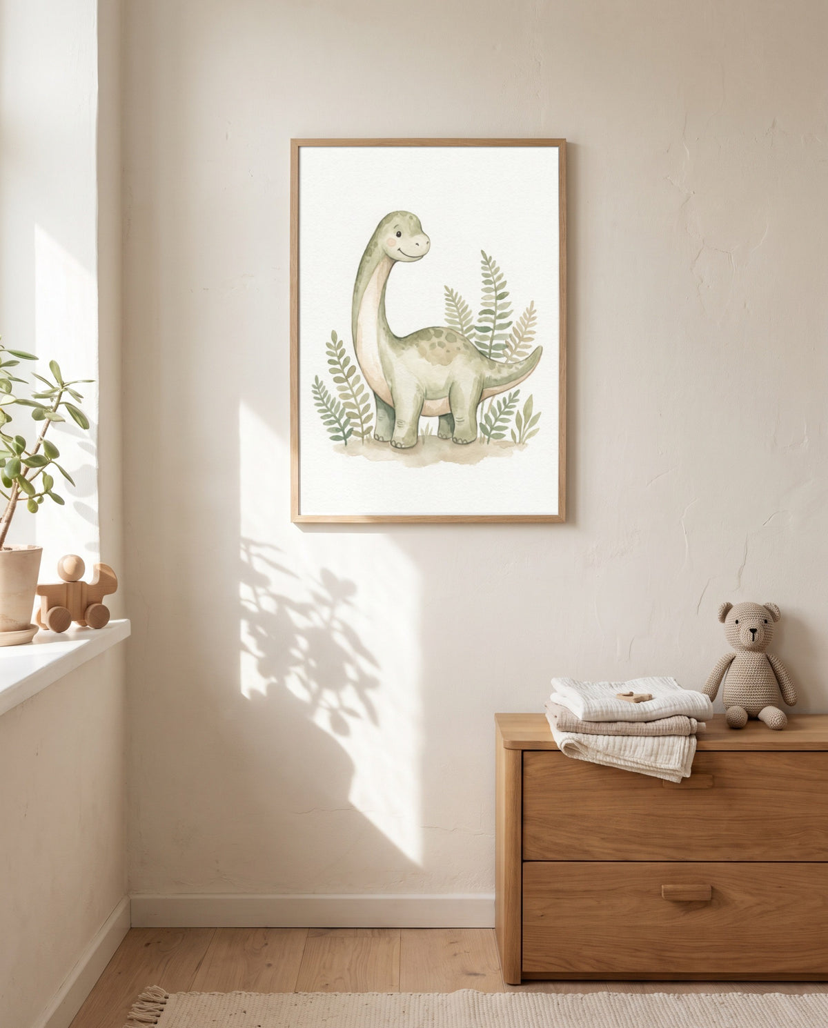 Langhals-Dino-Poster im hellen Holzrahmen an einer sonnenbeschienenen Wand über einer Holzkommode mit gefalteten Textilien und Stoffbär; am Fenster stehen eine Pflanze und ein Holzspielzeug.