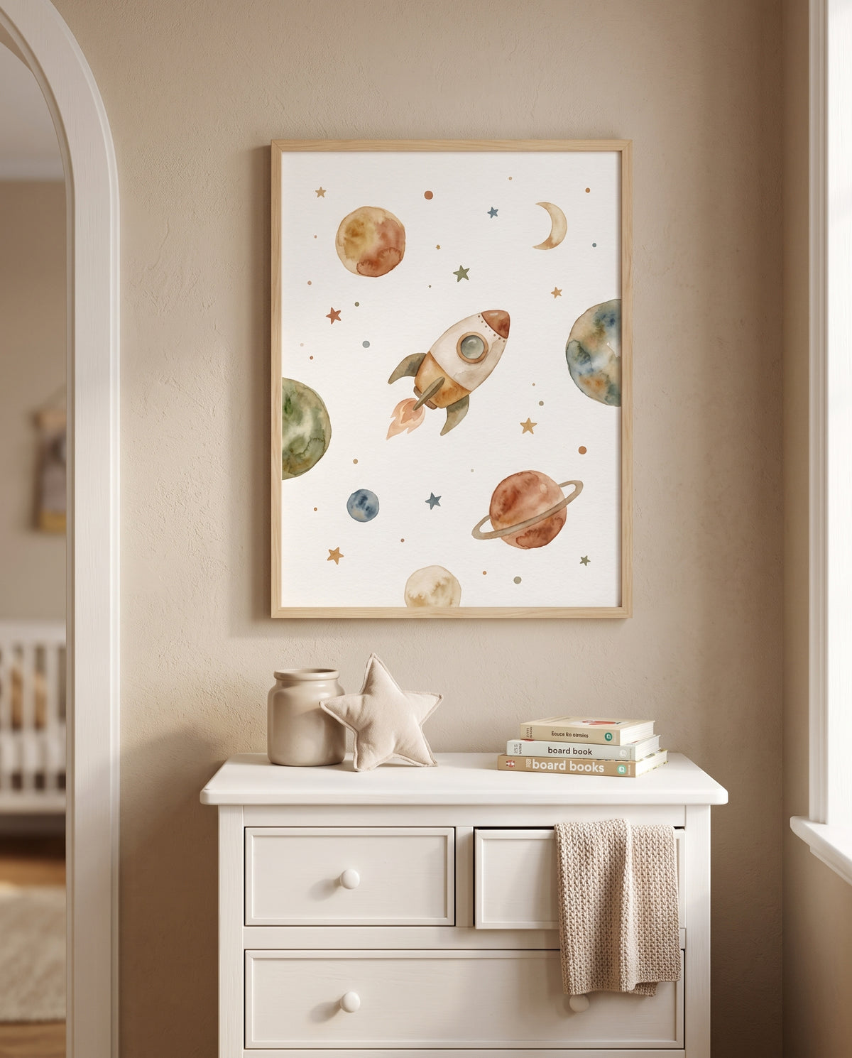 Weltraumposter mit Rakete und Planeten im Holzrahmen über einer weißen Kommode mit Gefäß, Stoffstern, Büchern und überhängendem Stricktuch, zwischen einem Rundbogen und einem Fenster.